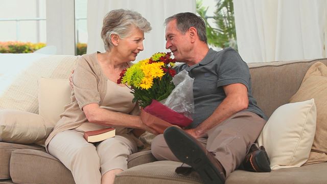 Elderly couple sitting closely on a couch holding a colorful bouquet, sharing an intimate and affectionate moment. The cozy indoor environment suggests a warm, loving home. Perfect for campaigns focusing on senior living, retirement communities, romantic illustrations for couples in later life, or depicting familial warmth and love.