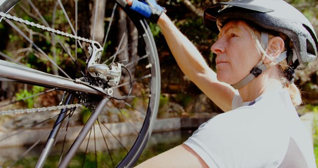 This image of a woman inspecting and maintaining a bicycle, suited up with a helmet, in an outdoor setting is perfect for promoting outdoor activities, biking, nature exploration, fitness, and healthy lifestyles. It can be used in blogs, articles, advertisements, or educational materials related to cycling, fitness tips, or women's empowerment in sports.