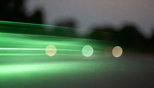 Gliding emerald light streaks sweeping low across pavement at dusk with three bokeh orbs