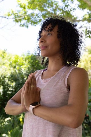 African american woman practicing meditation in sunlit park wearing smartwatch and pink tank