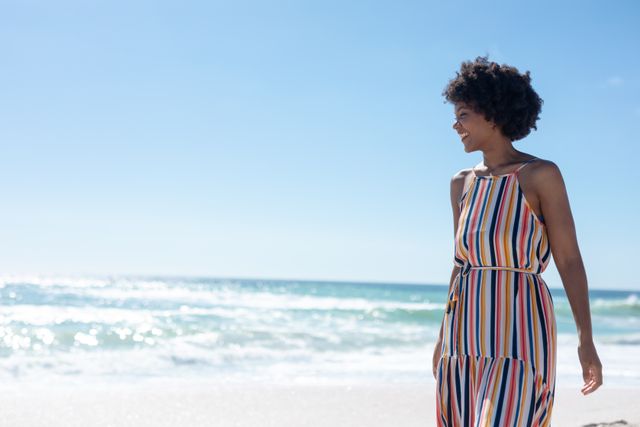Happy african american young woman strolling at beach by copy space against blue sky on sunny day. unaltered, people, lifestyle, enjoyment and holiday concept.