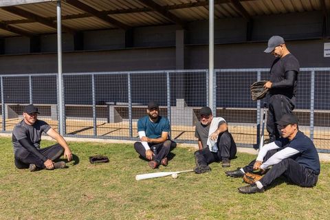 Diverse adult baseball teammates resting on grass in front of dugout with gloves and bat