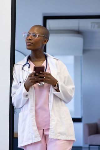 African woman clinician standing in doorway holding smartphone wearing lab coat and stethoscope