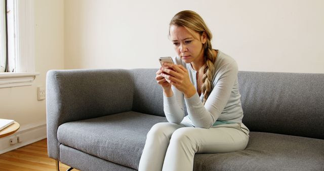 Woman sitting on a grey couch in a living room, looking concerned while using a smartphone. This could be used to depict situations involving worry, technology use, or personal moments in a home setting. Suitable for blogs, articles, or advertisements related to mental health, technology use, or home life.