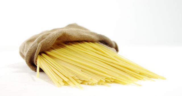Uncooked spaghetti noodles spill out from a burlap sack onto a white surface, with copy space. Spaghetti is a staple in Italian cuisine and is often used in a variety of pasta dishes.