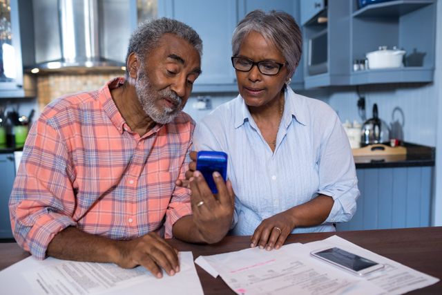 Senior couple sitting at kitchen table using calculator while discussing financial documents. Ideal for content related to retirement planning, budgeting, financial advice for seniors, and home finance management.