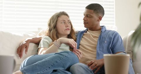 Interracial couple cuddling on sofa holding hands in sunlit living room