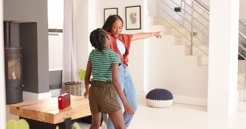 African mother and daughter dancing and laughing in sunlit modern living room near staircase