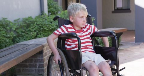 Blond boy sitting in wheelchair near building entrance with wooden bench and courtyard