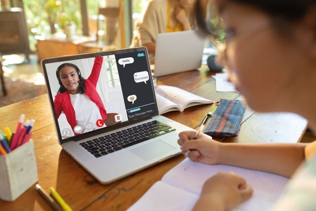 Asian teenage girl with book looking at laptop screen with student raising hand in online class. Home, video call, unaltered, internet, wireless technology, education, student and e-learning concept.