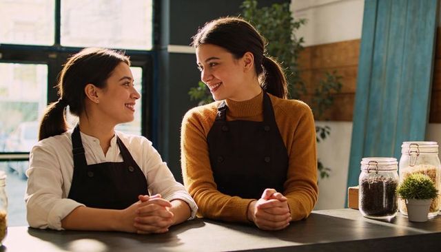 Two baristas smiling and chatting behind counter in sunlit modern cafe with glass jars
