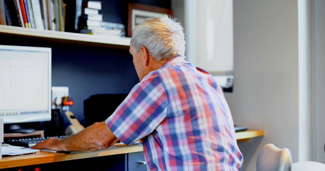A senior Caucasian man is focused on working at his computer in a home office environment, with copy space. His experience reflects the growing trend of older adults engaging with technology and remaining active in the workforce.