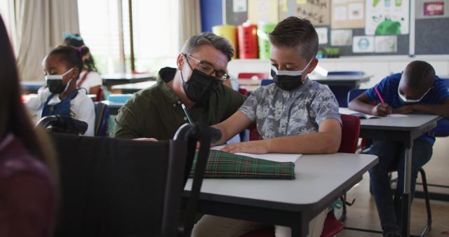Diverse male teacher helping schoolboy sitting in classroom, all wearing face masks. children in primary school during coronavirus covid 19 pandemic.