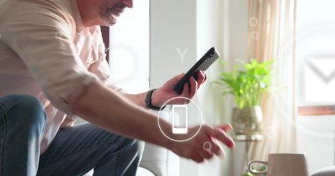 Senior man holding smartphone reaching for coffee cup on sofa with floating smart icons