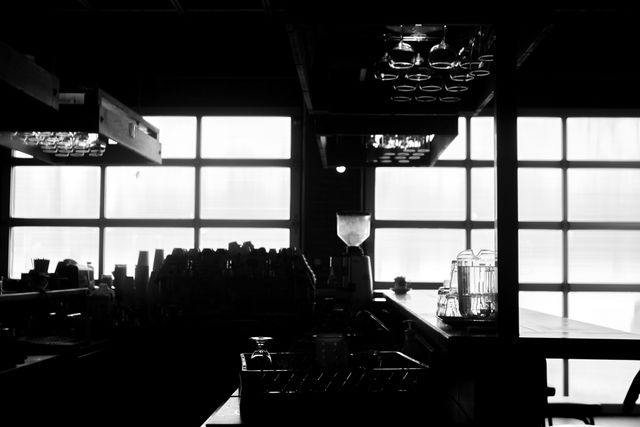 This dark silhouette of a bar interior, backlit by a large window, showcases an industrial, minimalist design. Ideal for use in publications and media highlighting modern bar designs, nightlife atmospheres, or interesting use of light and shadow.