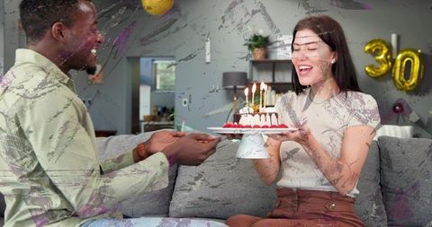 Couple celebrating 30th birthday on sofa with cake with lit candles and gold 30 balloons