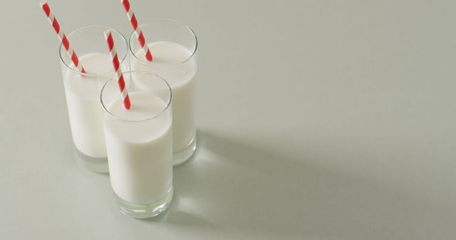 Three glasses of fresh milk are placed on a light surface, each with a red and white striped straw. The minimalist arrangement and clean background create a sense of simplicity and freshness. Ideal for promoting dairy products, healthy living, nutrition campaigns, and breakfast ideas.