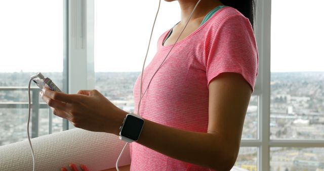 Woman wearing pink shirt holding yoga mat and smartphone while wearing smartwatch. Ideal for themes of modern fitness, technology in exercise, indoor activities, or healthy living. Suitable for promoting fitness apps, wearables, or home workout routines.