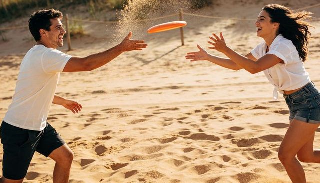 Laughing couple tossing orange frisbee across sunlit sandy dune on summer beach