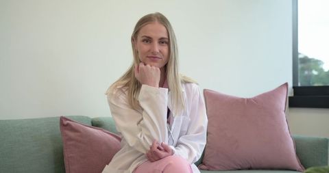 Medical professional sitting on pastel clinic sofa wearing white lab coat and pink scrubs