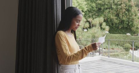 African American woman reading letter at doorway wearing mustard top and white jeans, balcony view