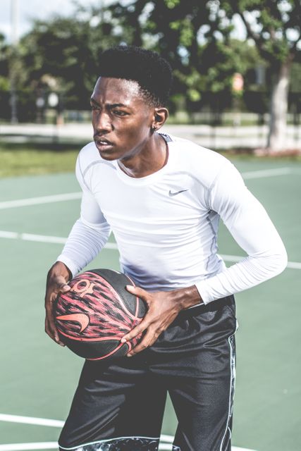 Young African American man wearing sportswear holding basketball on outdoor court. He appears determined and focused, perfect representation of athletic dedication. Useful for promoting sports gear, fitness programs, basketball events, and inspirational themes for physical activity.