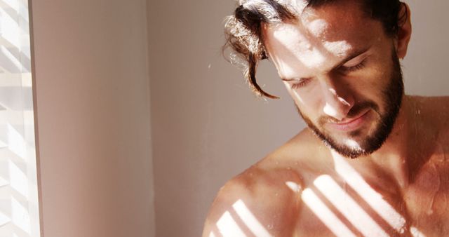 Young man with a well-groomed beard stands alone indoors, with sunlight casting dramatic shadows on his face and body. Natural light illuminates his casual, contemplative expression. Perfect for themes of self-confidence, masculinity, personal reflection, and modern lifestyle. Can be used in magazines, advertisements for grooming products, or health and wellness blogs focusing on men's mental health and self-awareness.