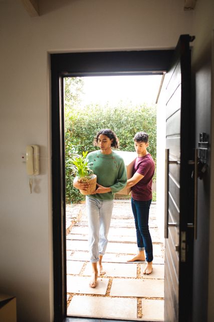 Multiracial young gay couple carrying potted plant and cardboard box seen through doorway. Copy space, moving, unaltered, love, togetherness, homosexual and new house concept.