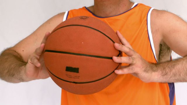 Detailed shot of a man dressed in an orange jersey holding a basketball, emphasizing his focus and physical movement. Useful for sports articles, training manuals, or athletic events promotions highlighting focus and agility.