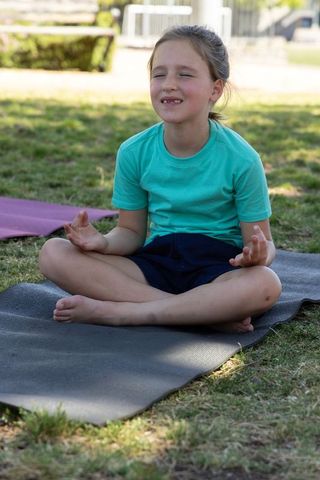 School-age girl meditating on yoga mat in park practicing mindful breathing