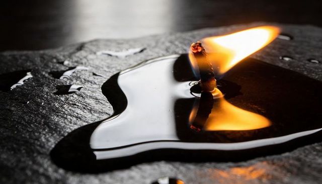Macro charred wick burning in oil pool on dark slate reflecting golden flame