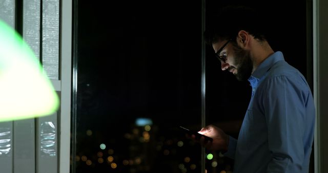 Man wearing blue shirt using smartphone by office window at night. City lights visible in background. Ideal for concepts related to business, technology, working late, professional communication, and night time work.