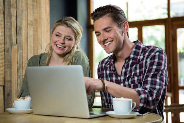 Young couple sitting at a coffee shop table, smiling and using a laptop. Ideal for illustrating themes of teamwork, collaboration, modern lifestyle, and casual work environments. Suitable for use in advertisements, blog posts, and social media content related to technology, relationships, and coffee culture.