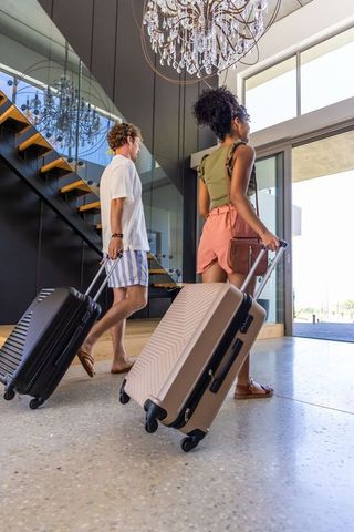 Multiracial couple walking through modern hotel lobby pulling rolling luggage under chandelier