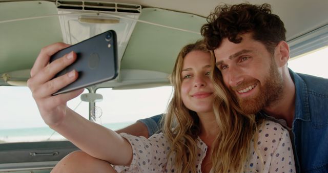 A cheerful couple captures a happy moment by taking a selfie in a camper van. It appears they are enjoying a road trip or vacation by the beach during the summer. Useful for advertising travel, tourism, adventure, and happy lifestyle content.