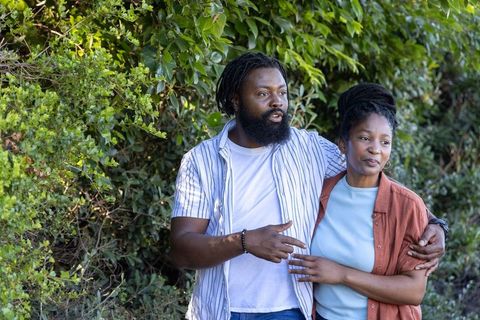 African couple walking by leafy hedge, man gesturing while woman smiles and leans