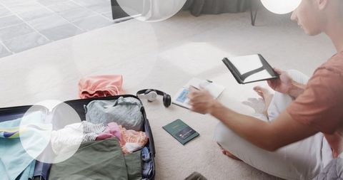 Man packing suitcase and organizing passport, tablet and travel essentials at home