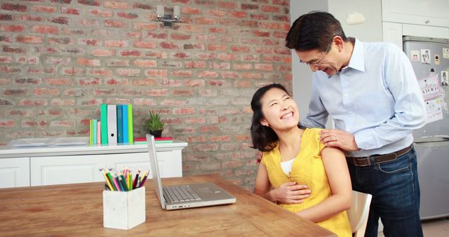 An Asian middle-aged man is standing next to a young woman seated at a wooden table with a laptop, both smiling in a room with a brick wall background. Their interaction suggests a positive and supportive relationship, in a home or casual office environment.