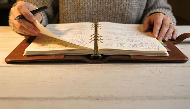 Turning Page in Leather Planner with Handwritten Notes and Pen on Wooden Desk