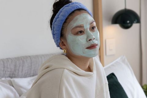 Asian woman wearing green facial mask and blue headband relaxing on bed in cozy bedroom