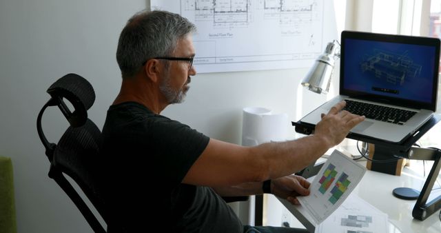 Senior architect reviewing detailed building plans on both a laptop and printed paper while sitting at a modern office workstation. Ideal for use in content related to architecture, engineering, construction management, project planning, and professional workspace environments.