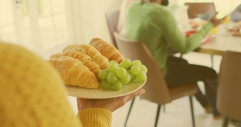 Senior in yellow knit serving croissants and green grapes at sunlit family breakfast