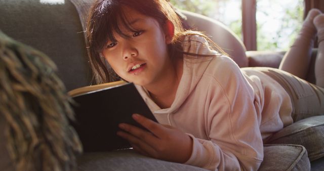 Asian girl smiling while reading a book while lying on the couch at home. childhood, hobby and leisure activity concept