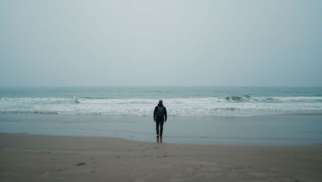 Solitary traveler facing foggy ocean at empty shoreline wearing backpack and beanie