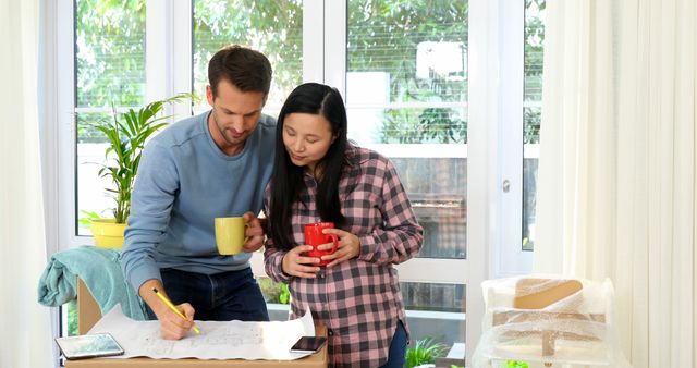 Young Caucasian man and Asian woman review plans at home. They're engaged in a home improvement project, sharing ideas over coffee.