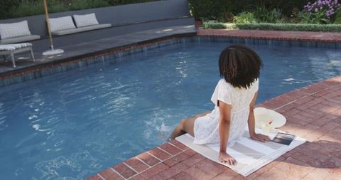 African american woman sitting by pool, dipping foot in water on brick deck, relaxing