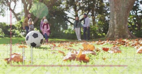 Soccer ball sitting on autumn lawn with fallen leaves and family walking toward ball in park