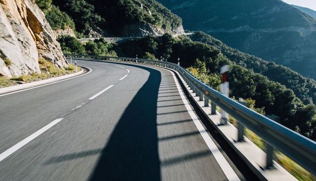 Winding mountain road curving along rocky cliff with guardrail and dramatic shadows