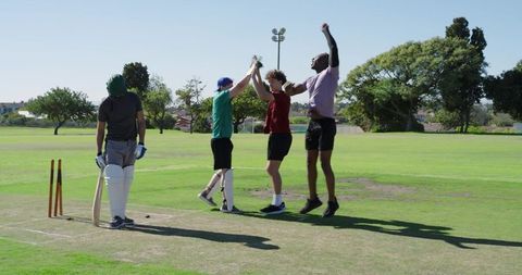Young cricketers celebrating wicket with high fives on sunny park pitch showcasing teamwork