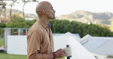 Mature man standing on balcony holding coffee mug gazing at morning landscape and horizon